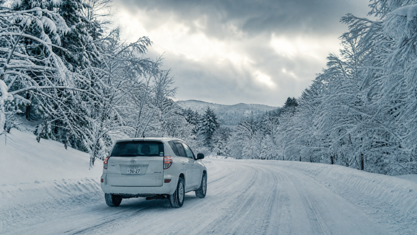 White SUV driving on a snow-covered road flanked by snow-laden trees on a cloudy winter day.