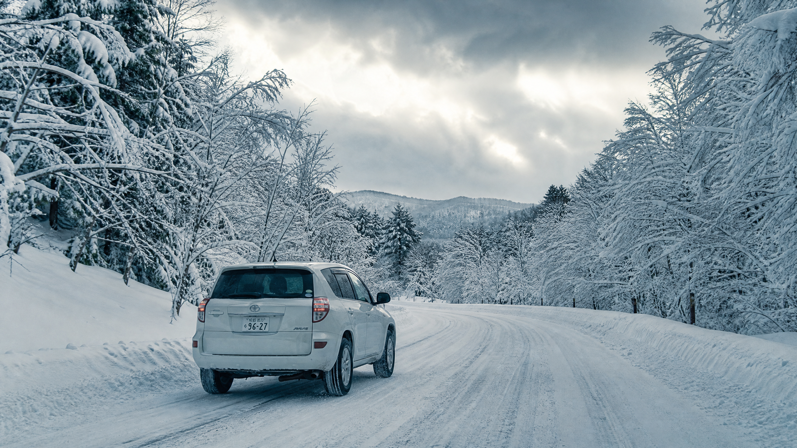White SUV driving on a snow-covered road flanked by snow-laden trees on a cloudy winter day.