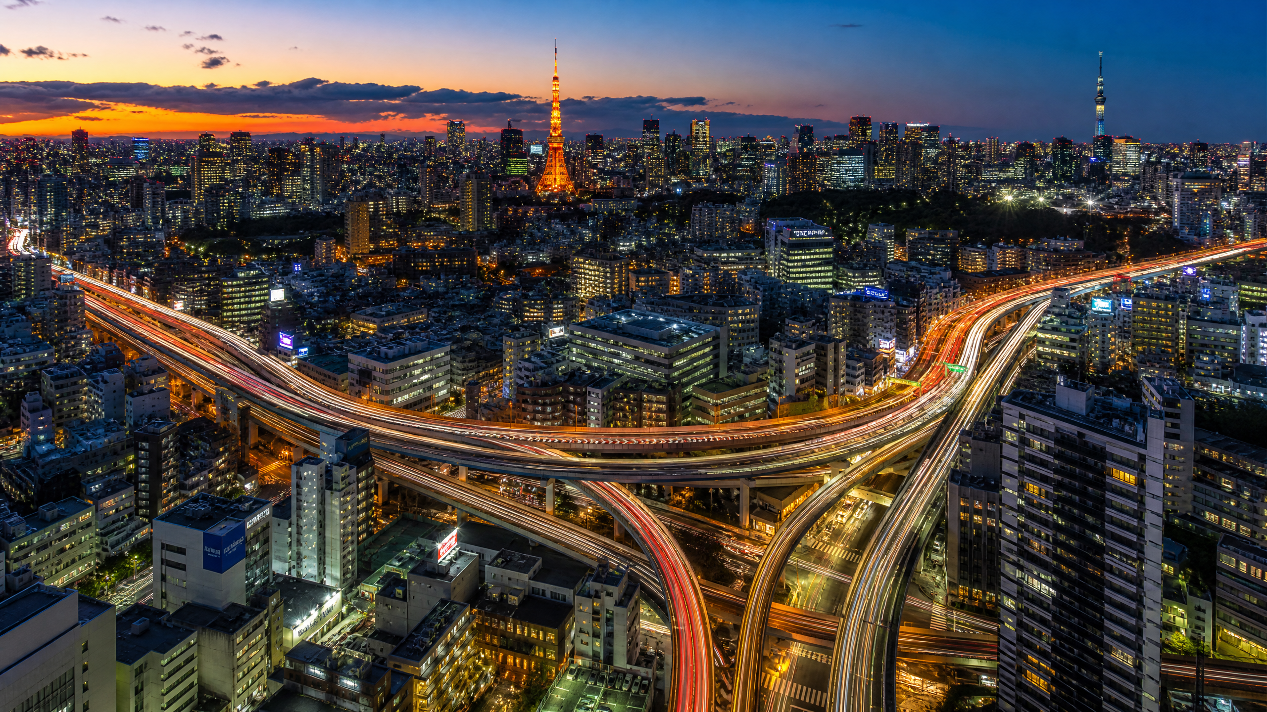 Aerial night view of Tokyo with the illuminated Skytree and winding freeway ramps glowing red and white across the cityscape at dusk.