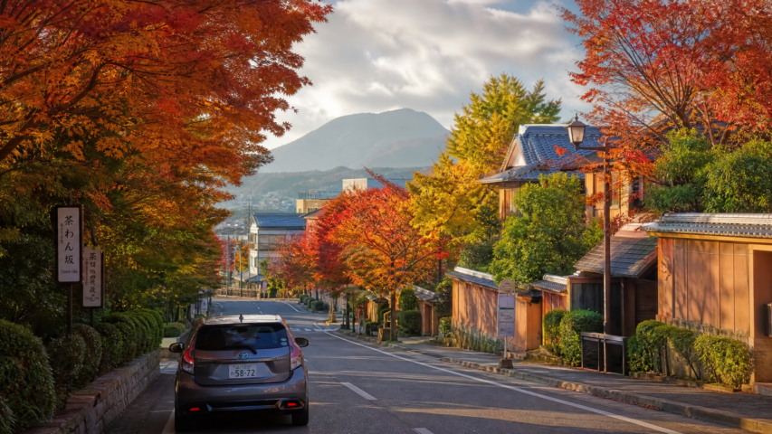 Street in a Japanese town during autumn, with red and orange maple trees lining both sides, traditional houses on the right, and a distant mountain.