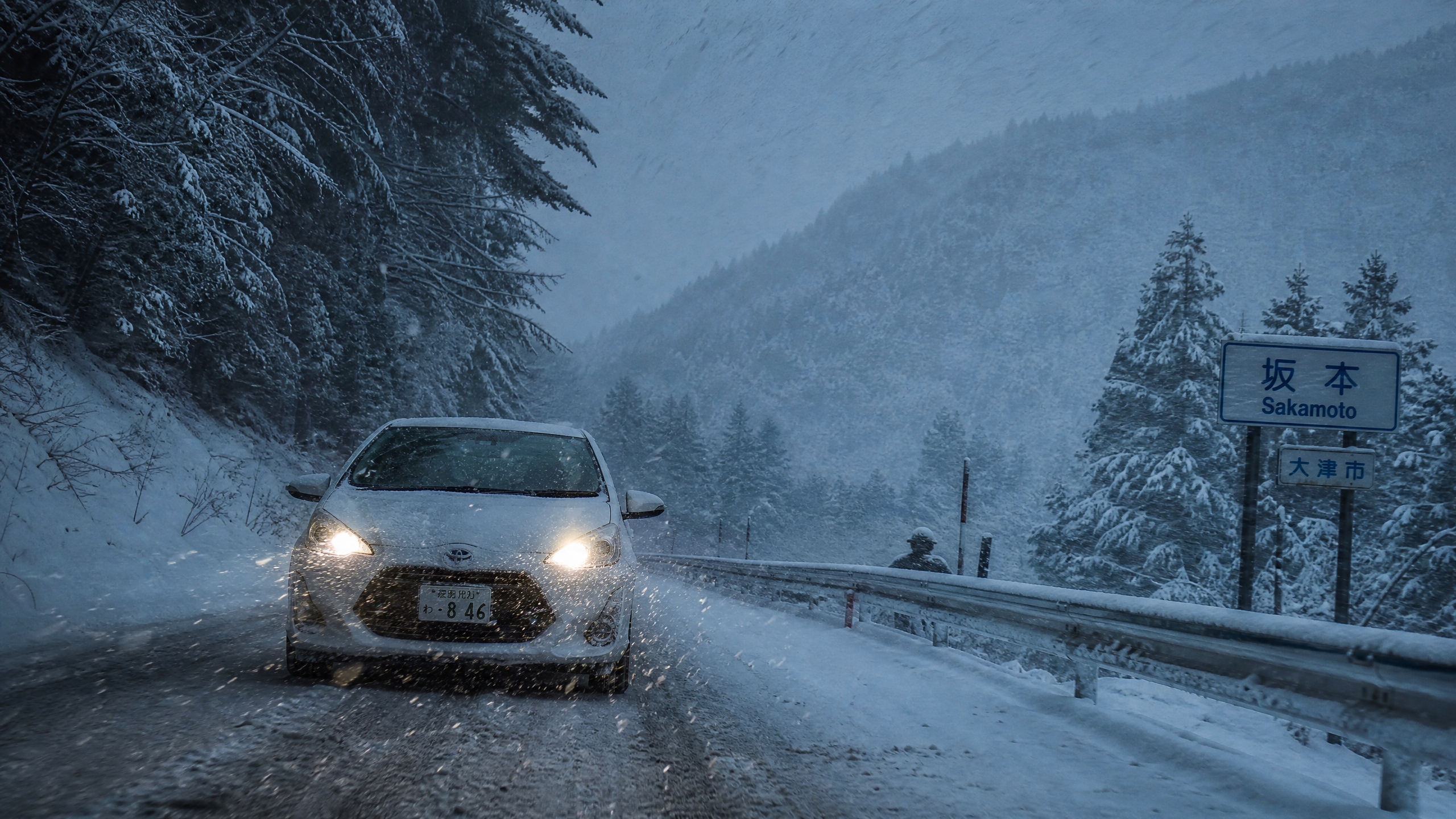 White car driving toward the camera on a snow-covered mountain road, headlights on, with snow-laden trees and a sign reading 'Sakamoto' in Japanese on the right.