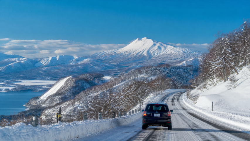 Snowy mountain road winding beside a frozen lake, with a car in the foreground and snow-covered trees under a clear blue sky.