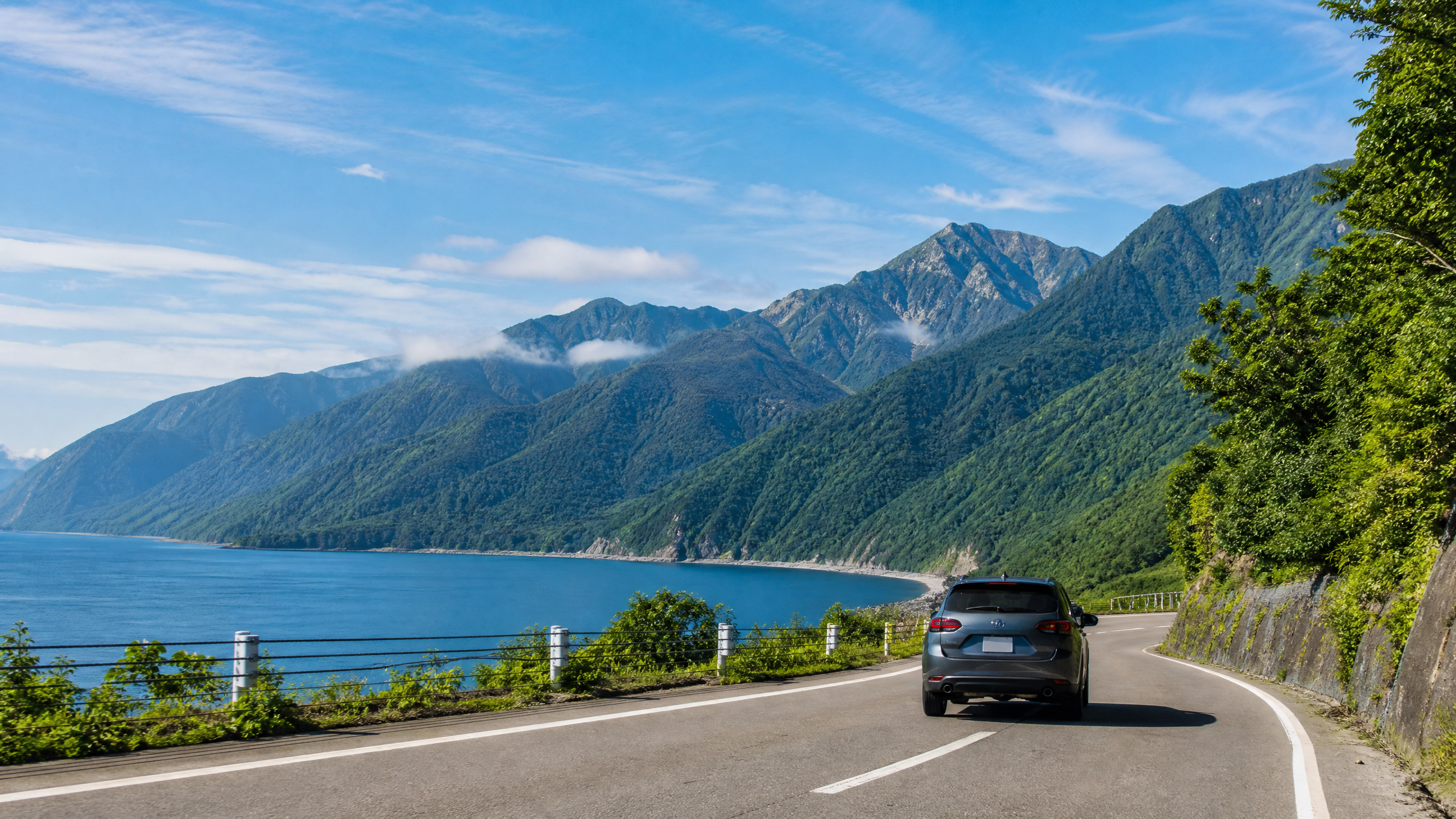 Car driving along a coastal mountain road with blue sea on the left and green hills on the right under a clear sky