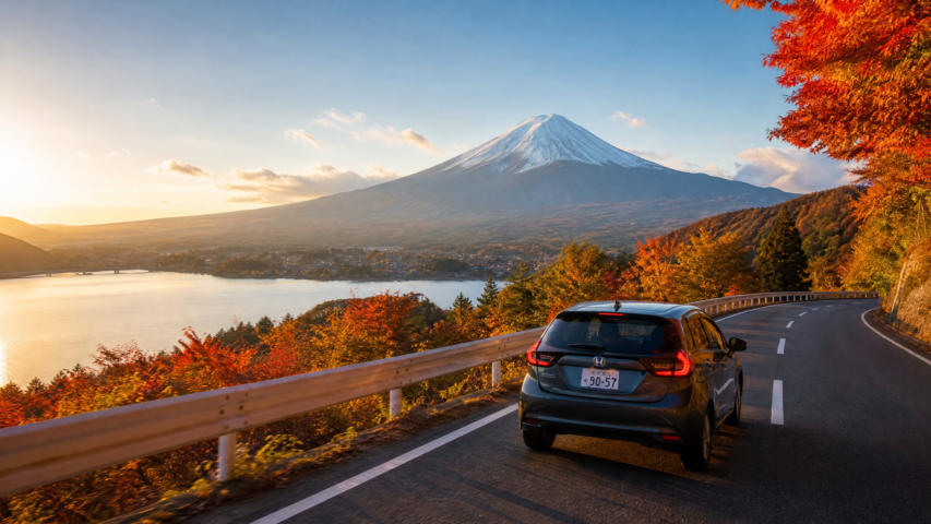 Car on a winding hillside road at sunset, autumn trees in orange and red, with a snow-capped mountain in the distance (Mt. Fuji).