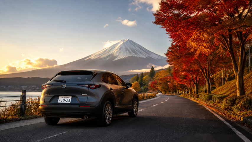 Rear three-quarter view of a gray Mazda SUV parked along a seaside road, with autumn red trees and a snow-capped mountain in the distance at sunset.
