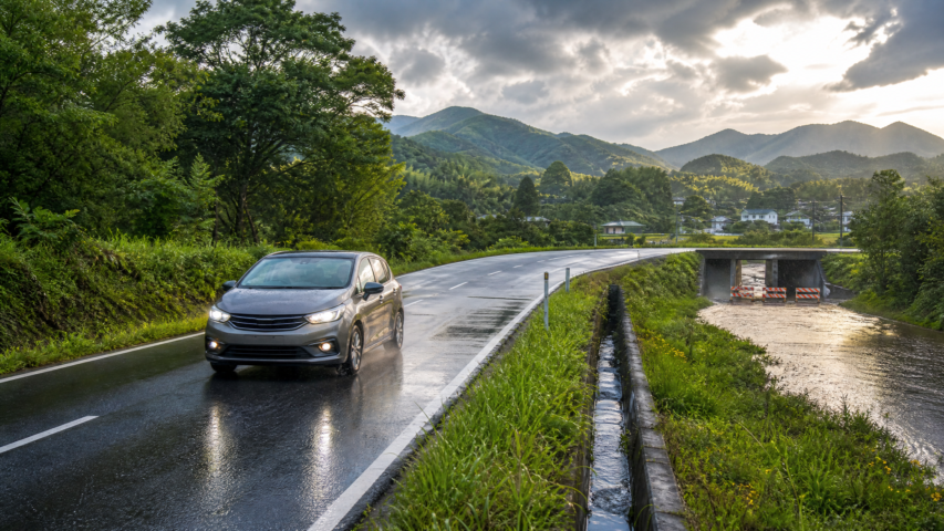 Gray sedan driving on a wet rural road beside a grassy canal, with green hills and a cloudy sky in the distance.