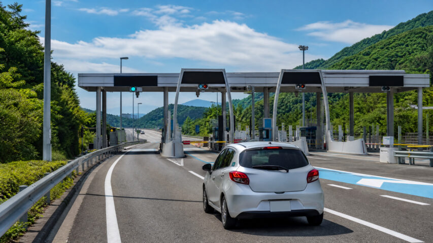 Silver car approaching a toll plaza with multiple lanes and green hills in the background, on a sunny day with a blue sky.