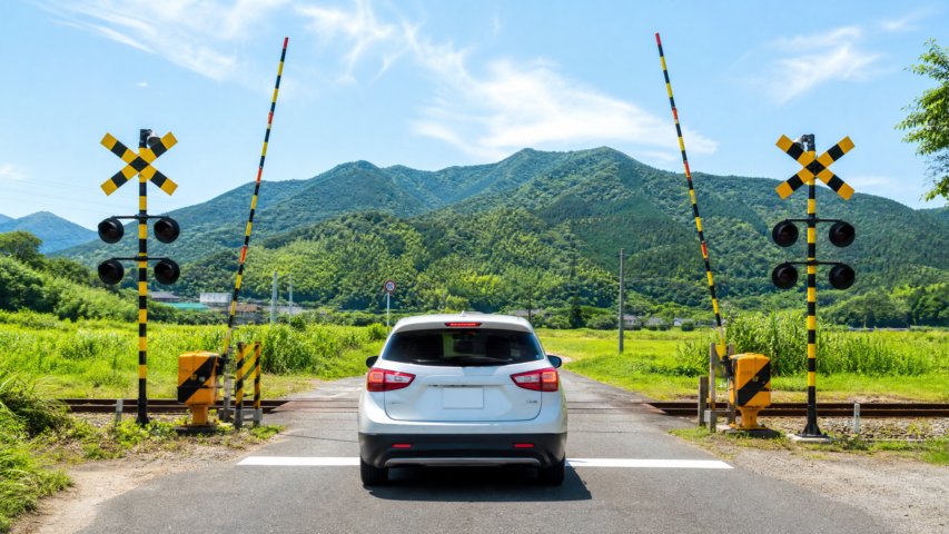 White SUV waiting at a rural railroad crossing with yellow-black striped gates and signal lights, green fields and blue mountains in the distance.