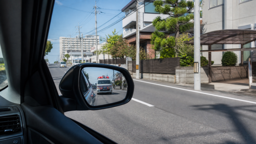 Side-view mirror shows a street scene with an ambulance directly behind the car on a sunny day.