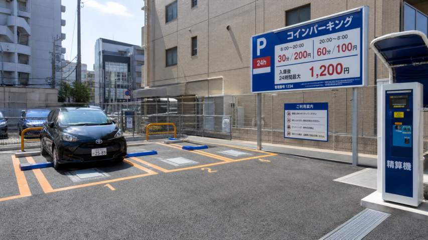 Black Toyota parked in a 24h coin parking lot with orange-lined spaces and a large blue-and-white rate sign nearby to the right gym-like kiosk