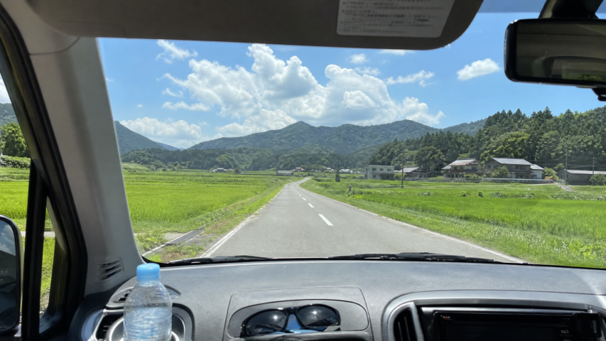View from inside a car driving down a rural two-lane road, with green fields and distant hills under a blue sky with clouds.