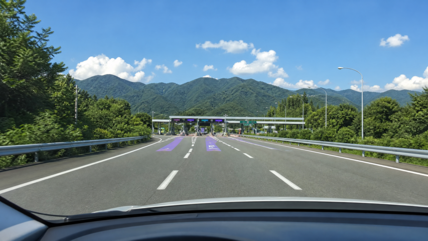 Driver’s-eye view on a multi-lane highway heading toward toll booths, with purple lane markings and green forested mountains under a blue sky in the distance