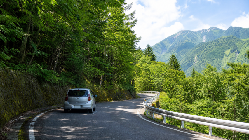 Curvy mountain road winding through a forest, with a silver car on the left and distant green mountains under a blue sky.