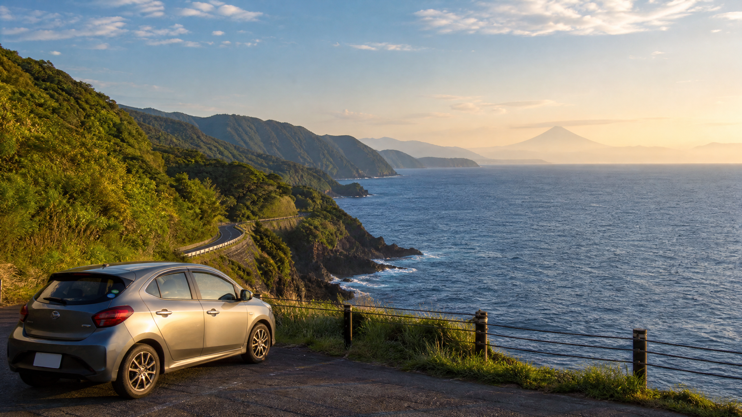 Silver hatchback parked on a coastal road overlooking the sea at sunset, with green hills and distant mountains.