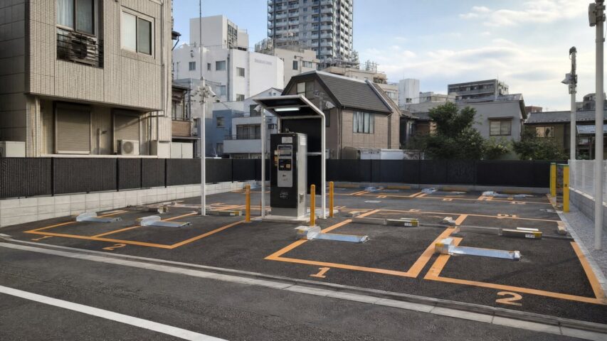 Outdoor urban parking lot with orange-lined spaces and a central kiosk under a small shelter, surrounded by buildings.