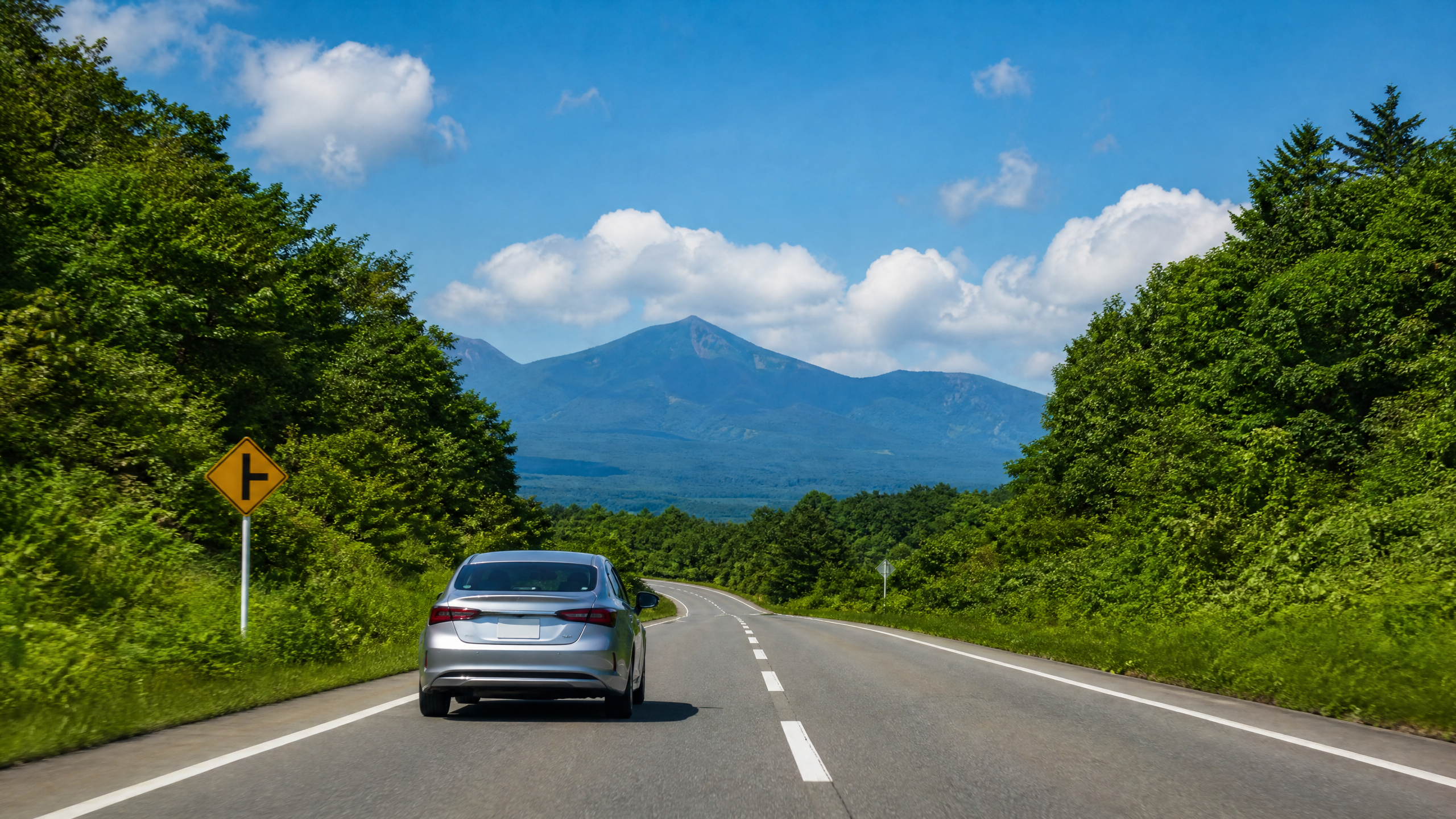 Silver sedan driving on a winding two-lane road with green trees on both sides and distant mountains under a blue sky.