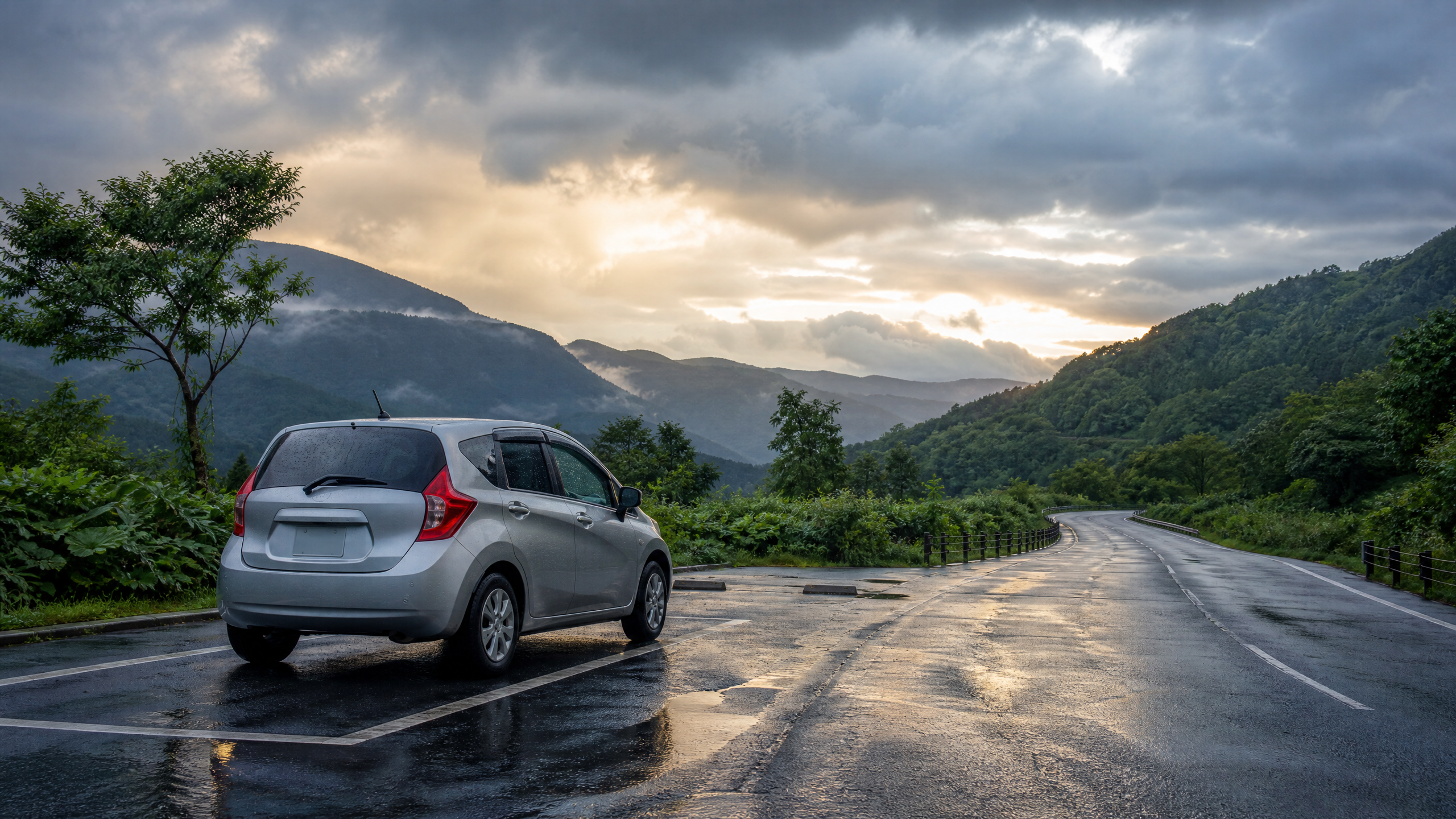 Silver hatchback parked on a wet mountain road with reflections on the asphalt and green hills under a cloudy sky at sunset/rainlight.