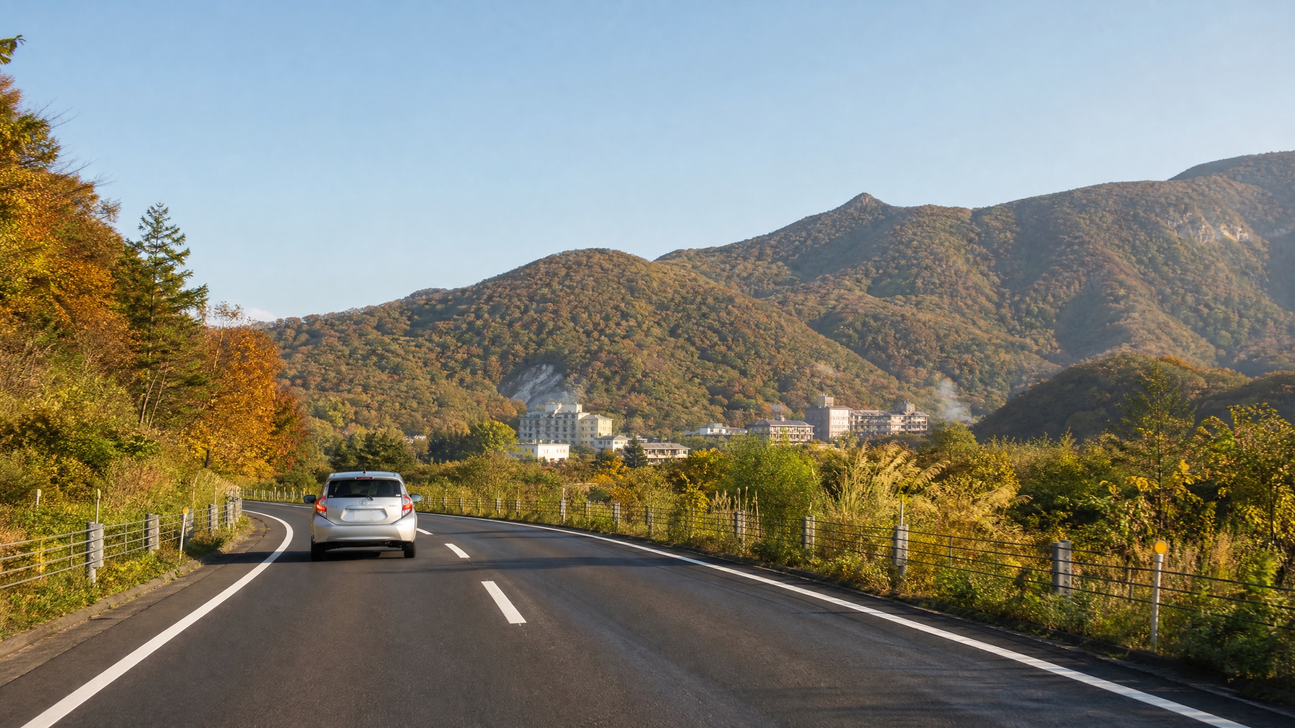 A calm early-autumn rental-car drive in Hokkaido toward a forested hot-spring town